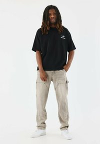 Camiseta negra de gran tamaño con un logo blanco, combinada con pantalones cargo gris claro. Zapatillas blancas completan el look casual.