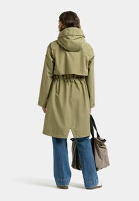 Wodoodporny trencz w oliwkowej zieleni o krótkim kroju, z taliowaniem i kapturem. Połączony z szerokimi niebieskimi dżinsami i szarą torbą typu tote.