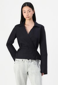 Black wrap shirt with long sleeves, collar, and tie waist detail, paired with light striped jeans. Smooth fabric with structured fit.