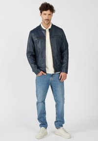 Marineblaue Lederjacke mit Stehkragen, mit Reißverschluss versehenen Taschen, kombiniert mit hellblauen Jeans und weißen Sneakers. Glatte Textur, moderner Schnitt.