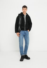 Schwarze, mit Fleece gefütterte Jacke mit einem Kragen aus Kunstleder, Reißverschluss und Akzentnähten, kombiniert mit hellblauen Jeans und schwarzen Stiefeln.