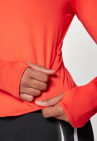 Langärmliges Sportoberteil aus leuchtend orangefarbigem Stoff, mit Daumenlöchern in den Bündchen, glatter Textur und taillierter Passform.