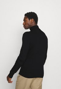 Black ribbed turtleneck sweater, fitted design with long sleeves, soft texture, and a high neckline, paired with light-colored pants.