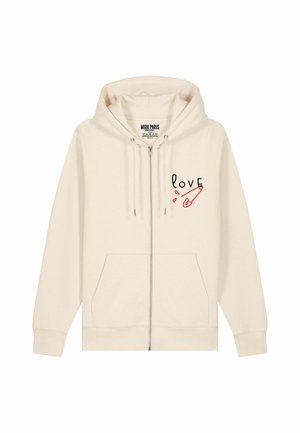 Haut à zip crème en tissu doux. Il dispose de poches avant et de l'inscription "love" accompagnée d'un graphique de épingle de sûreté rouge sur la poitrine gauche.