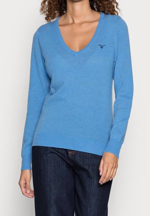 Kvinde iført en himmelblå V-hals langærmet sweater og mørkeblå jeans, stående mod en ensfarvet baggrund.