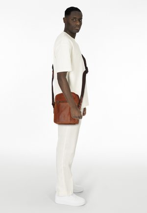 Borsa a tracolla in pelle marrone con forma rettangolare, tracolla scura regolabile e tasca frontale con zip, mostrata su un modello che indossa un abbigliamento bianco.