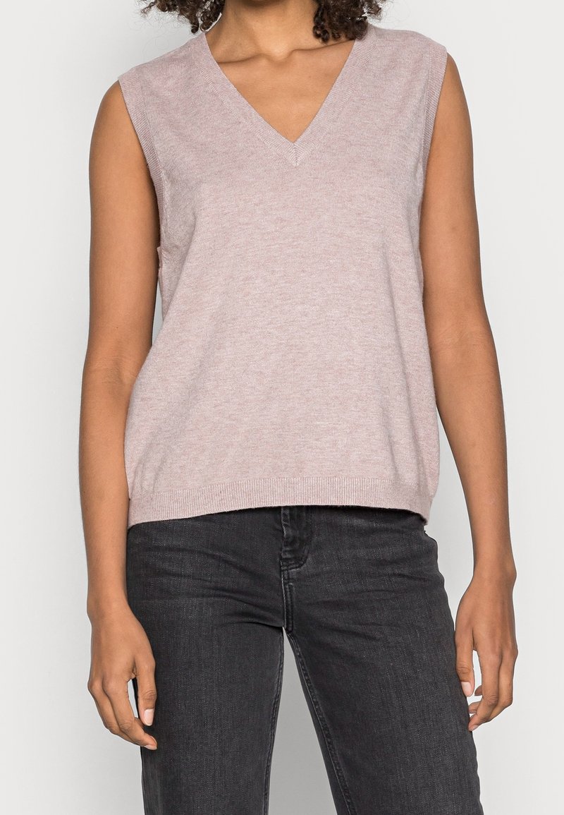 Haut en maille sans manches, couleur mauve clair, col en V, porté avec un jean gris foncé sur une personne debout devant un fond uni.