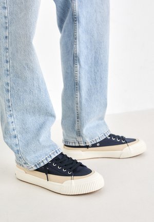 Tengerészeti kék és krémszínű, alacsony szárú sportcipők, érdes gumitalppal és fűzős elülső résszel, világoskék egyenes szárú farmernadrággal párosítva.