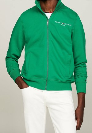 Mężczyzna w zielonej kurtce na suwak z logo "Tommy Hilfiger" i białych spodniach, jedna ręka w kieszeni, na jednolitym tle.