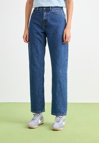 Jean droit taille haute bleu clair en denim, avec deux poches avant et des coutures contrastées, associé à des baskets grises.