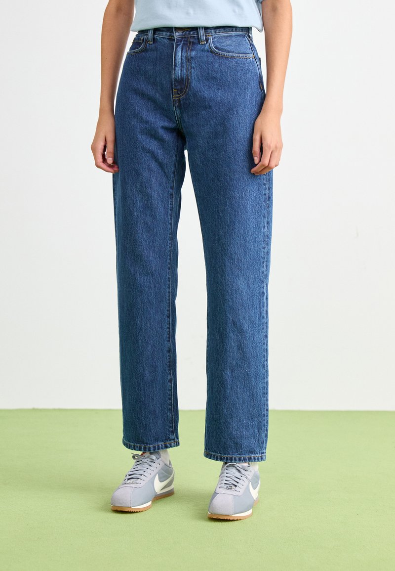 Jean droit taille haute bleu clair en denim, avec deux poches avant et des coutures contrastées, associé à des baskets grises.