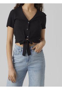 Top corto nero con maniche corte, scollo a V, bottoni frontali e orlo annodato, abbinato a jeans a vita alta azzurri. Texture del tessuto liscia.