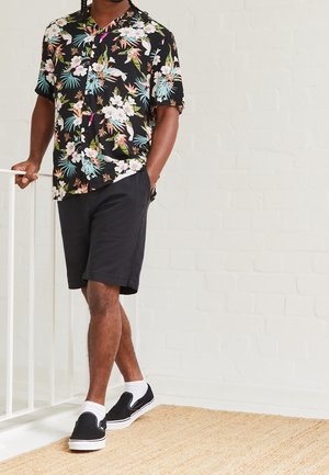 Man wearing black floral short-sleeve shirt, black shorts, white socks, and black slip-on shoes leaning on white railing indoors.