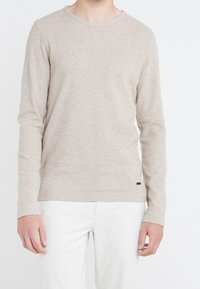 Person klädd i en beige långärmad tröja med rund hals och vita byxor mot en slät vit bakgrund.