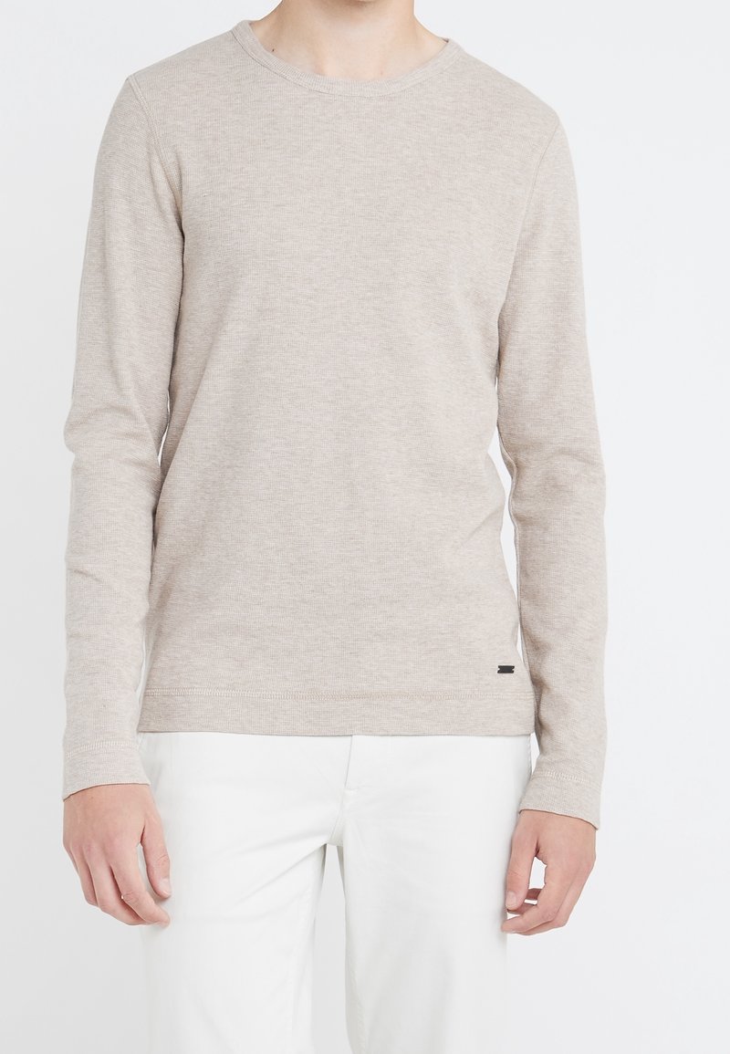 Person klädd i en beige långärmad tröja med rund hals och vita byxor mot en slät vit bakgrund.