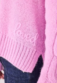 Rosa gestrickter Pullover mit einer weichen, flauschigen Textur. Das Wort "geliebt" ist in einem leicht dunkleren Rosa gestickt. Gerippter Saum und Ärmel.