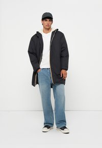 Doudoune noire avec capuche, dotée d'une fermeture éclair et d'accents jaunes. Associée à un jean bleu clair et des baskets noires.
