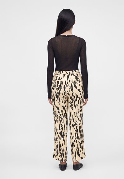Femme aux longs cheveux noirs portant un haut noir transparent à manches longues et un pantalon beige à jambes larges avec des taches abstraites noires, debout de dos.