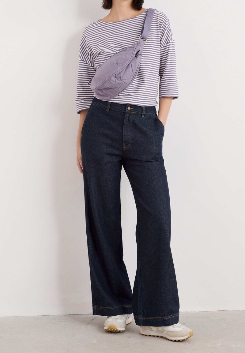 Personne portant un jean large foncé, des baskets blanches, un haut à manches trois-quarts rayé et un sac bandoulière lavande sur un fond uni.