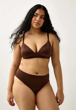 Kvinde med langt krøllet sort hår iført brun bh og høj talje undertøj, stående mod en ensfarvet lys baggrund.