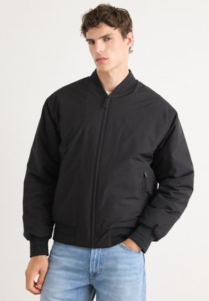 Zwarte bomberjack met een rits aan de voorkant, ribgebreide manchetten en zoom. Voorzien van een zijzak en een gladde, matte textuur. Draag het over lichtblauwe jeans.