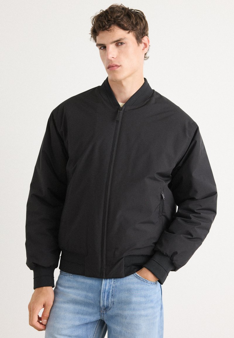 Black bomber jacket with a zip front, ribbed cuffs, and hem. Features a side pocket and a smooth, matte texture. Worn over light blue jeans.