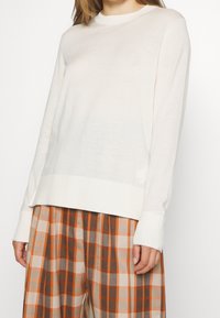 Bílý svetr s dlouhým rukávem a kulatým výstřihem, s žebrovaným lemem a manžetami, spárovaný s béžovými a oranžovými kostkovanými kalhotami.