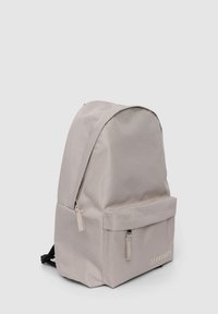 Zaino in tessuto grigio con forma arrotondata, tasca frontale, zip in metallo e branding discreto sul pannello frontale inferiore.