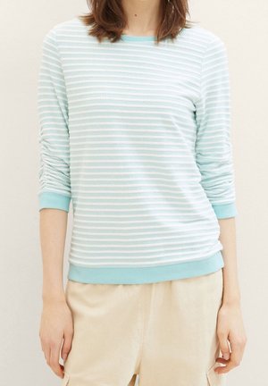 Vrouw draagt een lichtblauw-wit gestreept shirt met lange mouwen met geplooide mouwen en beige broek, staand tegen een effen achtergrond.