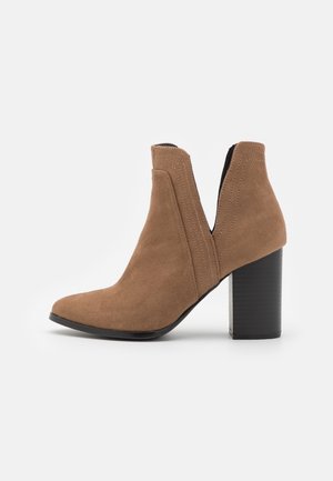 Bottine en suede marron avec un bout pointu, des côtés découpés et un talon épais noir. Des accents de couture apportent des détails au design.