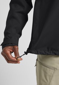 Schwarze wasserdichte Jacke mit verstellbarem Kordelzug am Saum, mit glatter Textur und einer Nahaufnahme einer Hand, die am Kordelzug zieht.