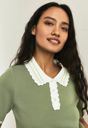 Young woman with long dark hair wearing a green knit top with white scalloped collar and pearl buttons, smiling softly against a plain background.