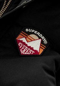 Crni materijal sadrži pravougaonu izvezenu oznaku s dizajnom planine, suncem i natpisom "SUPERDRY EVERST" u bijeloj i crvenoj boji.