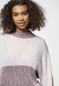 Maglione a maglia con texture a costine, corpo rosa chiaro e una fascia viola scuro attorno al collo e alla sezione centrale.