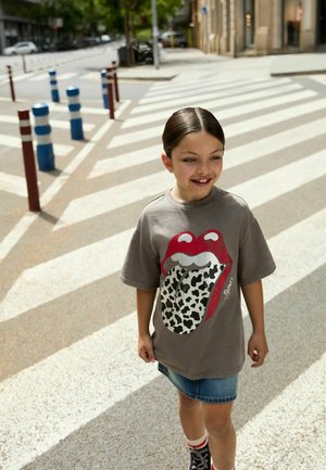 Junges Mädchen trägt ein graues T-Shirt mit Aufdruck und einen Jeansrock und läuft über einen großen gestreiften Zebrastreifen in einer Stadtstraße.
