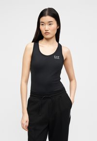 Νεαρή γυναίκα με μακριά σκούρα μαλλιά, ντυμένη με μαύρο αμάνικο τοπ EA7 Emporio Armani και μαύρο παντελόνι, στέκεται με το ένα χέρι στην τσέπη.