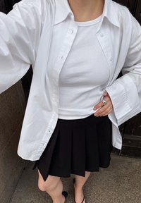 White button-up shirt over a white ribbed tank top, paired with a black pleated skirt. The skirt features a curved hem and is knee-length.