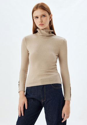 Beige lange mouwtop met een hoge gerimpelde kraag, gladde textuur, getailleerd ontwerp en knoopaccenten op de polsen. Gecombineerd met donkere jeans.