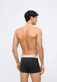 Mužský model zobrazený zozadu, oblečený v čiernych vzorovaných boxerách Calvin Klein s bielym pásom, stojaci na svetlom jednoduchom pozadí.