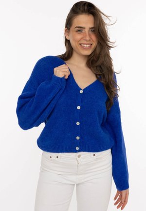 Frau lächelt, trägt eine kobaltblaue, geknöpfte Strickjacke und weiße Hose, steht vor einem schlichten weißen Hintergrund.