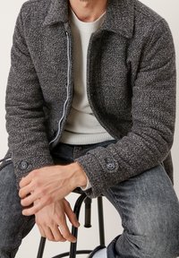 Chaqueta gris con textura, con cuello, cierre de cremallera y puños con botones, llevada sobre un suéter claro, combinada con jeans oscuros.