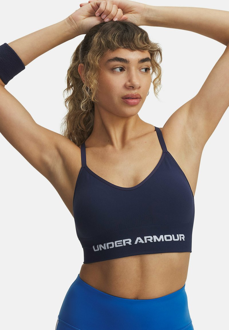 Marineblauwe sportbeha met dunne bandjes, een V-halsontwerp en een brede witte "UNDER ARMOUR" logo op de band. Gladde stofstructuur.