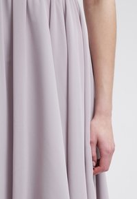 Gros plan du bras et de la main d'une personne à côté d'une robe fluide lavande clair avec de doux plis.