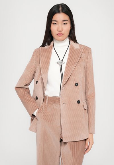 Frau trägt beigen Cord-Double-Breasted-Blazer und dazu passende Hose mit weißem Rollkragenpullover und schwarzem Bolo-Tie-Anhänger.