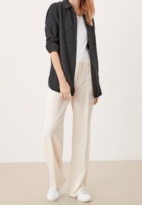 Chemise noire à boutons, en tissu de lin, manches longues retroussées, associée à un pantalon large en lin de couleur crème et des baskets blanches.