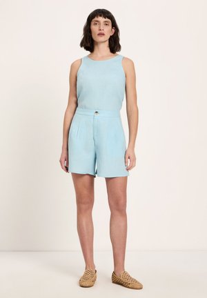 Frau steht, trägt ärmelloses hellblaues Oberteil und passende hoch taillierte Shorts mit gewebten hellbraunen flachen Schuhen vor einfachem Hintergrund.