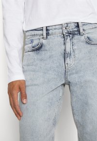 Ljust blå denimjeans med en lätt urblekt finish, fem fickor och kontrasterande gula sömmar. Matchas med en vit långärmad skjorta.