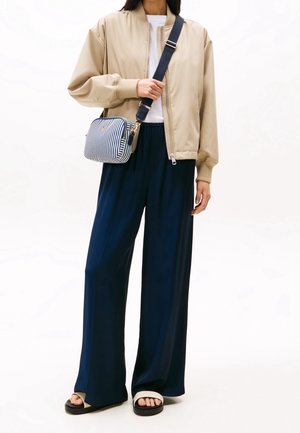 Beige bomberjakke med ribbede manchetter, marineblå vide bukser, sorte sandaler med hvide accenter og en stribet crossbody taske.