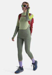 Vrouw in groene sportkleding, paarse sokken, blauwe wandelschoenen, rode rugzak en spiegelende zonnebril die haar bril aanpast, staand op een witte achtergrond.