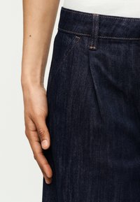 Χέρι που ακουμπά δίπλα σε σκούρα μπλε τζιν παντελόνι με ορατή ραφή και λεπτομέρεια μπροστινής τσέπης, μπροστά σε ανοιχτό φόντο.